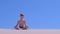 Child boy is meditating in lotus pose on sandy beach on blue sky background.