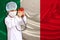 Child, boy, in a white doctorâ€™s coat, hat and mask attached a stethoscope to a red heart model, Italy flag background, close-up