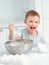 Child cooks food. Caucasian boy sits at table in kitchen and kneads dough in cup with hand whisk. Cheerful and positive