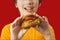 Child eats burger on red background. Male child with hamburger