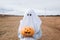 Child in ghost costume to halloween holding carved pumpkin in autumn field.