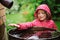 Child girl in red raincoat playing with water barrel in rainy summer garden. Water economy and nature care