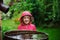 Child girl in red raincoat playing with water barrel in rainy summer garden. Water economy and nature care