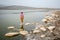 A child girl in a striped T-shirt and a yellow skirt is standing on a stone in the water in the lake.