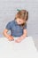 Child, a girl at the table writes, draws on a piece of paper, against a white brick wall