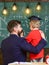 Child in graduate cap listening teacher, chalkboard on background, rear view. Teacher with beard, father hugs little son