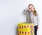 Child in gray jumper posing isolated on white. She leaning on yellow suitcase tied by white and red warning tape