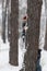Child hides behind tree in winter forest. Boy is dressed in warm hat and winter jumpsuit
