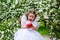 The child is holding a large red apple and wants to take a bite. A little happy girl in a dress plays under a blooming apple tree