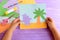 Child holds a paper card with a Hippo and a palm tree. Colored paper sheets, scissors, pencil, glue stick