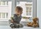 Child in home quarantine at the window putting a medical mask on his sick teddy bear, for protection against viruses during