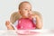 The child independently eats cottage cheese with a spoon from a pink plastic bowl, sitting on a chair