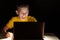 A child laughs out loud, sitting at a table in the dark and watching a video on a laptop screen