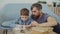 Child is learning, writing words in exercise book and his father is teaching him sitting near table and explaining rules