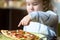 Child looks and points a finger at pizza on table. Three years old kid is going to eat by himself