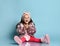 Child in pink faux fur coat, pants and boots. She smiling, gesticulating, sitting on floor against blue background. Close up