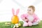 Child in pink fluffy costume sitting near yellow ostrich egg with bunny ears headband, colorful Easter eggs and tulips