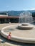 The child is playing next to the fountain. A little girl in a pink dress wets her hands near a large fountain in Budva