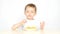 A child with pleasure eating potato chips and showing thumb of his hand sitting at a table on a white background