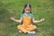 Child practices yoga on the street, sitting on green grass on summer day. Child is sitting in Lotus position