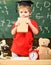 Child, pupil on cheerful face near microscope. Wunderkind concept. Kid boy in graduate cap holds book in classroom