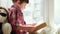 Child reading book, smart kid wearing checkered red shirt sitting on window and holding book