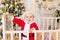 A child in a Santa costume stands in a crib at home near a Christmas tree with a Golden decor