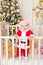 A child in a Santa costume stands in a crib at home near a Christmas tree with a Golden decor