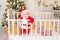 A child in a Santa costume stands in a crib at home near a Christmas tree with a Golden decor