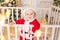 A child in a Santa costume stands in a crib at home near a Christmas tree with a Golden decor