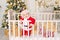 A child in a Santa costume stands in a crib at home near a Christmas tree with a Golden decor