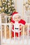 A child in a Santa costume stands in a crib at home near a Christmas tree with a Golden decor