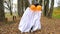 A child in sheets with cutout for eyes like a ghost costume dancing in an autumn forest scares and terrifies. A kind little funny