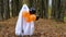 A child in sheets with cutout for eyes like a ghost costume dancing in an autumn forest scares and terrifies. A kind little funny