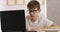 Child sits at table with laptop and does homework. Boy with glasses is studying online.
