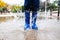 Child splashes in a puddle of water in a park with blue rain boots