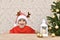 A child in a striped cap with deer horns laughs sitting at the table at the artificial Christmas tree and white lantern.