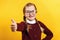 Child student with glasses shows thumb up. Little girl with pigtails in a burgundy sweater on a yellow background