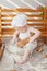 Child under one year old in a white cook apron and hat sits at a bench with baskets with rolls, bread, donuts, milk and flour