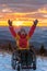 Child in wheelchair raising arms, enjoying sunset with majestic mountains in the background