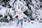 Child in white warm winter clothes and knit hat looking at the falling snow In winter forest