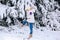 Child in white warm winter clothes and knit hat looking at the falling snow In winter forest