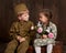 Children boy are dressed as soldier in retro military uniforms and girl in pink dress sitting on old suitcase, dark wood backgroun