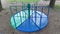 A children\\\'s carousel made of metal and wood is installed on the ground near the grassy lawn between the trees. The carousel and 