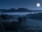 Chilling scenery of forested hills in fog with the full moon in the blue sky at night time