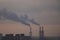 The chimneys of a refinery with smoke and steam with the pink and yellow sunset on the background