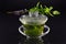 In a Chinese Gaiwan made of glass there is fresh warm peppermint tea against a dark background with fresh peppermint on the lid of