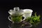 In a Chinese Gaiwan made of glass there is fresh warm peppermint tea in front of a dark background with a teacup, fresh peppermint