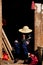 Chinese old women wearing traditional hat sits in wooden doors in morning light. She holding hat in hot weather. Yuanyang, Yunnan