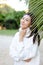 Chinese smiling girl in white sweater standing near palm leaf.